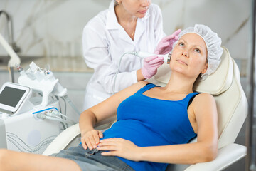 Young woman cosmetologist performs facial ultrasound procedure with machine to adult female client
