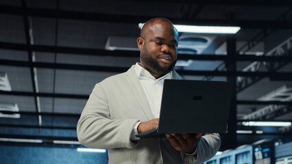 Engineer walking in server room backing up storage arrays using software on laptop. African...