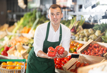 Man supermarket worker with vegetables in hands. He examines products, checks quality of some large tomatoes, rearranges products and forms showcase