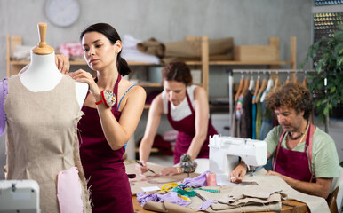 Creative female dressmaker working with mannequin while others sewing and drawing in tailor studio