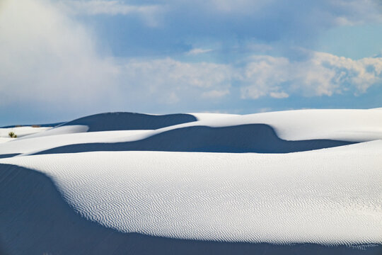 Rolling white sand dunes under blue cloudy skies in springtime with a rainstorm forming in the distance at White Sands National Park, New Mexico