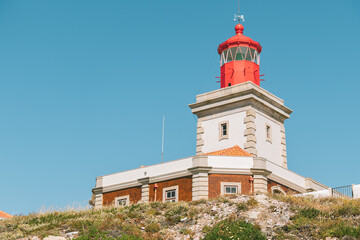 Bright red lighthouse stands on a rocky hill under clear blue sky in Lisbon region, Portugal. Ideal...