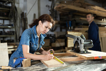 Woman employee of carpentry workshop makes measurements of wooden board, counts off set parameters...