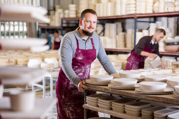 Young male worker in uniform moves rack with ceramic dishes blanks at factory