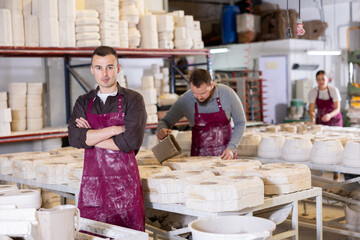 Confident ceramic artist in maroon apron standing with folded arms in pottery production workshop,...