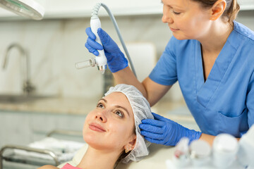Adult woman cosmetologist performs facial moisturizing procedure with machine to young female client