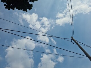Electric pole with power lines against a blue sky with clouds, showing urban energy infrastructure...