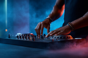 Hands actively adjusting controls on DJ controller in an atmospheric setting. Vibrant smoke and colorful lights create an energetic vibe. Concept of nightlife, entertainment, music events