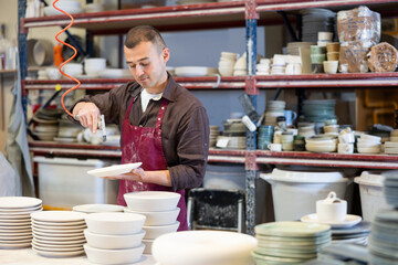 Skilled young adult male artisan in maroon apron working in pottery workshop, dusting off...