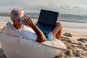 Mature man with laptop talking by mobile phone on beach. Business trip, watching video during preparing course work on laptop computer, sitting on the beach in sunny day. Happy work concept.