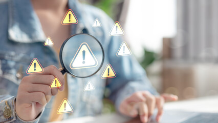 Close-up of person using magnifying glass to examine warning signs on virtual screen. Concept of cybersecurity alert, system error detection, risk management, and online safety monitoring.