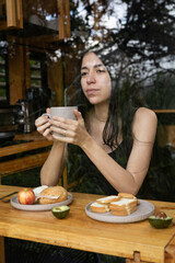 A front view of a young woman holding a coffee cup, seen from outside the window. The soft morning light and wooden textures of the glamping kitchen create a calm, peaceful mood.

