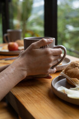 Artistic photo showing a hand holding a coffee cup on a breakfast bar, with blurred plates of eggs, fruit, and sandwich behind. Soft natural light creates a peaceful morning mood


