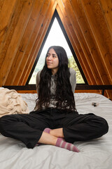A young woman sits thoughtfully on a cozy bed with her legs crossed and soles of her feet together, reflecting in a warm glamping tent filled with soft natural light.

