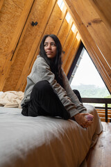 A young woman sits peacefully cross-legged in a lotus pose on a cozy bed inside a rustic glamping tent, bathed in soft natural light, embracing calm and mindfulness.

