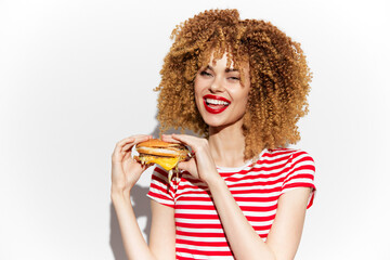 Happy woman enjoying a delicious burger, wearing a striped shirt, with curly hair and a bright...