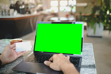 A person works on a laptop with a blank green screen while holding a white card, perfect for business or e-commerce mockups in a cafe setting.