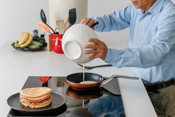 Elderly man pouring pancake batter into a frying pan in modern kitchen. Concept of homemade food,...