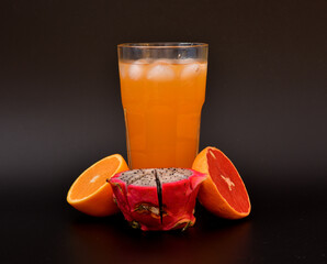 Tropical fruit juice in a tall glass with ice on a black background, next to it are pieces of ripe orange, grapefruit and pitaya.