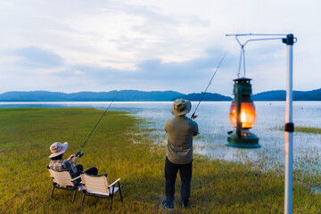 Romantic couple fishing together by the lake at dusk, sitting on camping chairs with lanterns and drinks nearby. Quiet outdoor lifestyle surrounded by nature.
