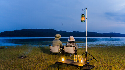 Romantic couple fishing together by the lake at dusk, sitting on camping chairs with lanterns and drinks nearby. Quiet outdoor lifestyle surrounded by nature. © wanakit