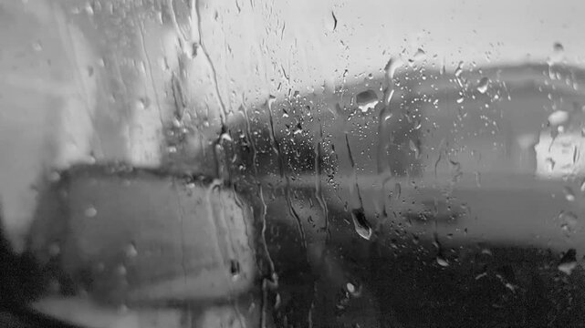 It's raining outside. Black and white video with wet glass. There's a car outside. Raindrops are spreading across the window.