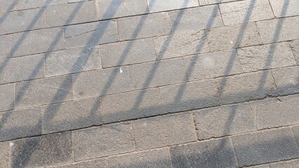Sunlight casts distinct, parallel shadows across a textured surface of gray rectangular paving...