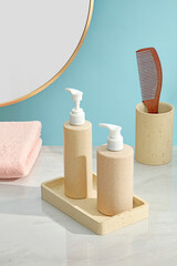 Neutral pump bottles on wooden tray beside comb, cup and mirror, against pastel blue wall. Neatly spaced composition offers room for cosmetic or label insert.