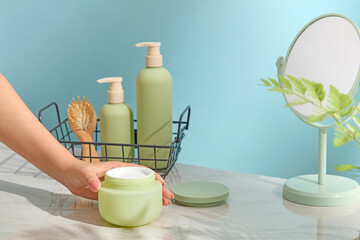 Hand holding body cream near pastel green skincare bottles and round mirror. Light blue background enhances wellness vibe. Ample space for product layout.