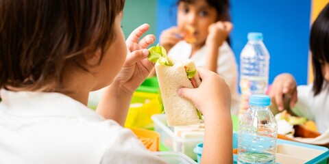 Children eating sandwiches at a table. Kids enjoy healthy sandwiches. Bottled water and lunchboxes...