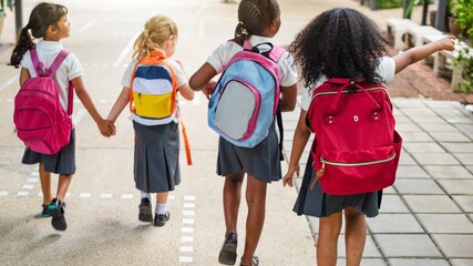 Four young girls with backpacks walk together on a path. They wear school uniforms and hold hands,...