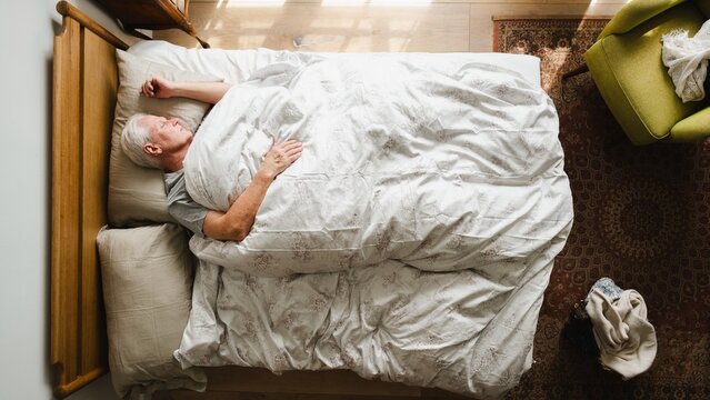 Elderly person sleeping in bed, covered with a white blanket. Peaceful rest, cozy bed, serene sleep. Top view of a calm, restful scene in a bedroom. Elderly man sleeping in bed, in retirement home. - Powered by Adobe
