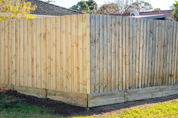 A timber boundary fence at the corner of a suburban residential property in Australia, showing a clear contrast between newly installed pale wood panels on left and weathered, aged timber on the right