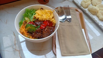 A delicious plate of black pepper barbecue beef served over steamed rice, accompanied by scrambled eggs and a side of fresh vegetable salad. A hearty and flavorful meal.