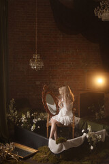 Young Caucasian woman with long blonde hair, in white dress, rear view, sits by a vintage mirror in a dark moody room with brick wall and flowers. Concepts: reflection, solitude, fantasy, dream.