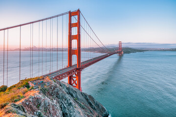 Golden Gate Bridge at sunrise in San Francisco, California. Scenic view from Battery Spencer with soft pastel sky, calm bay waters, and iconic red architecture. Tranquil travel landscape.