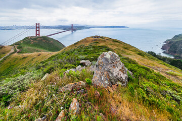 Panoramic view of the Golden Gate Bridge spanning San Francisco Bay with cityscape, lush hills, and boats, captured from a high vantage point on a cloudy spring day