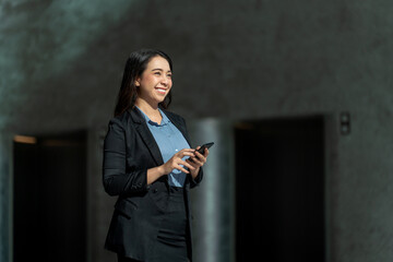 Smiling Asian businesswoman using smartphone in modern office lobby. Confident young professional...
