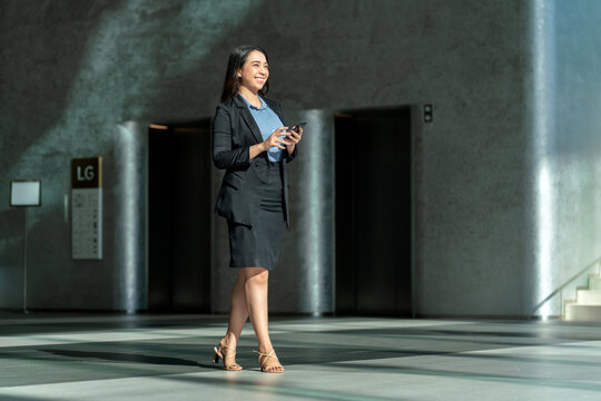 Smiling Asian businesswoman using smartphone in modern office lobby. Confident young professional walking with phone in elegant corporate interior
