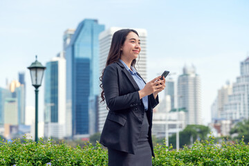 Successful Asian businesswoman using phone outdoors with skyscrapers in background. Young...