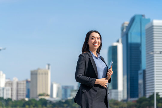 Smiling Asian businesswoman holding tablet with modern city skyline. Confident female professional outdoors in urban business district.