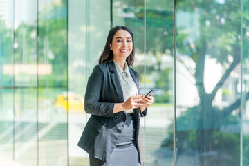 Asian businesswoman using smartphone near glass wall with reflection. Confident woman in formal outfit using phone in modern indoor space.