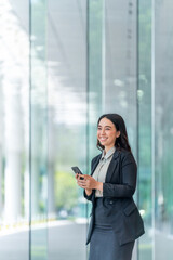 Fototapeta premium Smiling Asian businesswoman using smartphone in modern glass corridor. Confident woman in business attire standing indoors with mobile phone.
