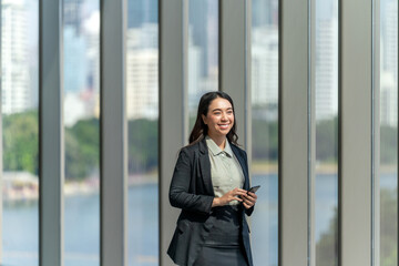 Fototapeta premium Asian businesswoman using smartphone indoors near panoramic window with city view. Confident professional woman in office hallway with natural light.