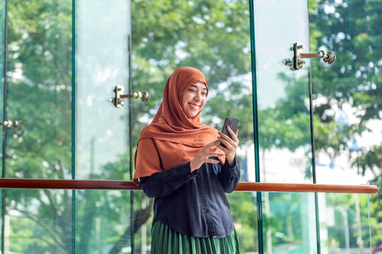 Asian muslimah using mobile phone with cheerful expression in bright modern space. Happy Muslim woman holding smartphone near glass window with greenery outside.
