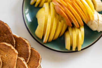 Close-up of healthy breakfast with sliced apple, banana, and pancakes on white table and green plate