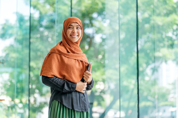 Smiling Asian muslimah holding tablet in bright modern office. Confident Muslim woman standing with...