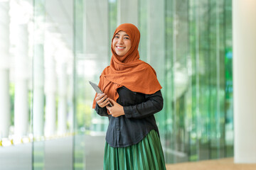 Smiling Asian muslimah holding tablet in bright modern office. Confident Muslim woman standing with...