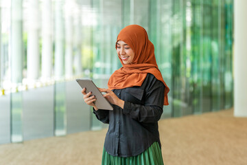 Asian muslimah using tablet near glass wall in bright modern building. Confident Muslim woman...