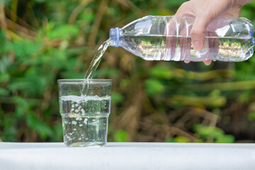 A hand pours clear, pure drinking water from a plastic bottle into a transparent glass, set against a blurred natural green background. The scene evokes freshness, hydration, and healthy living.